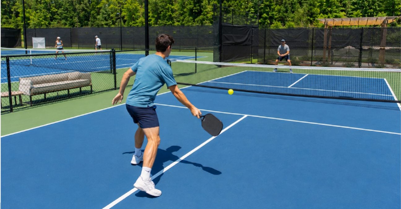 Tennis player on a pickleball court holding a paddle near the kitchen line with a tennis court behind