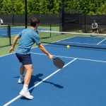 Tennis player on a pickleball court holding a paddle near the kitchen line with a tennis court behind