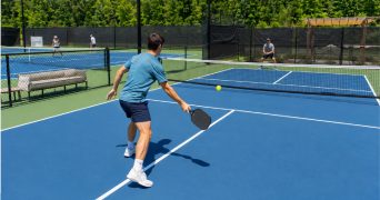 Tennis player on a pickleball court holding a paddle near the kitchen line with a tennis court behind