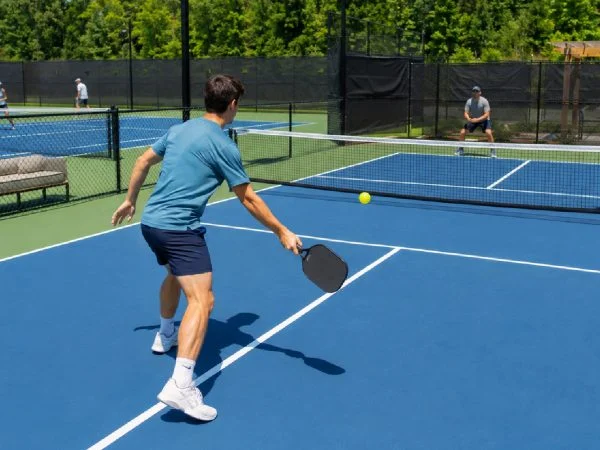 Tennis player on a pickleball court holding a paddle near the kitchen line with a tennis court behind